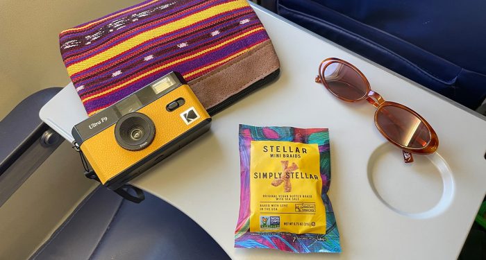 Food tray on plane with Stellar Snacks, camera, sunglasses, and travel bag