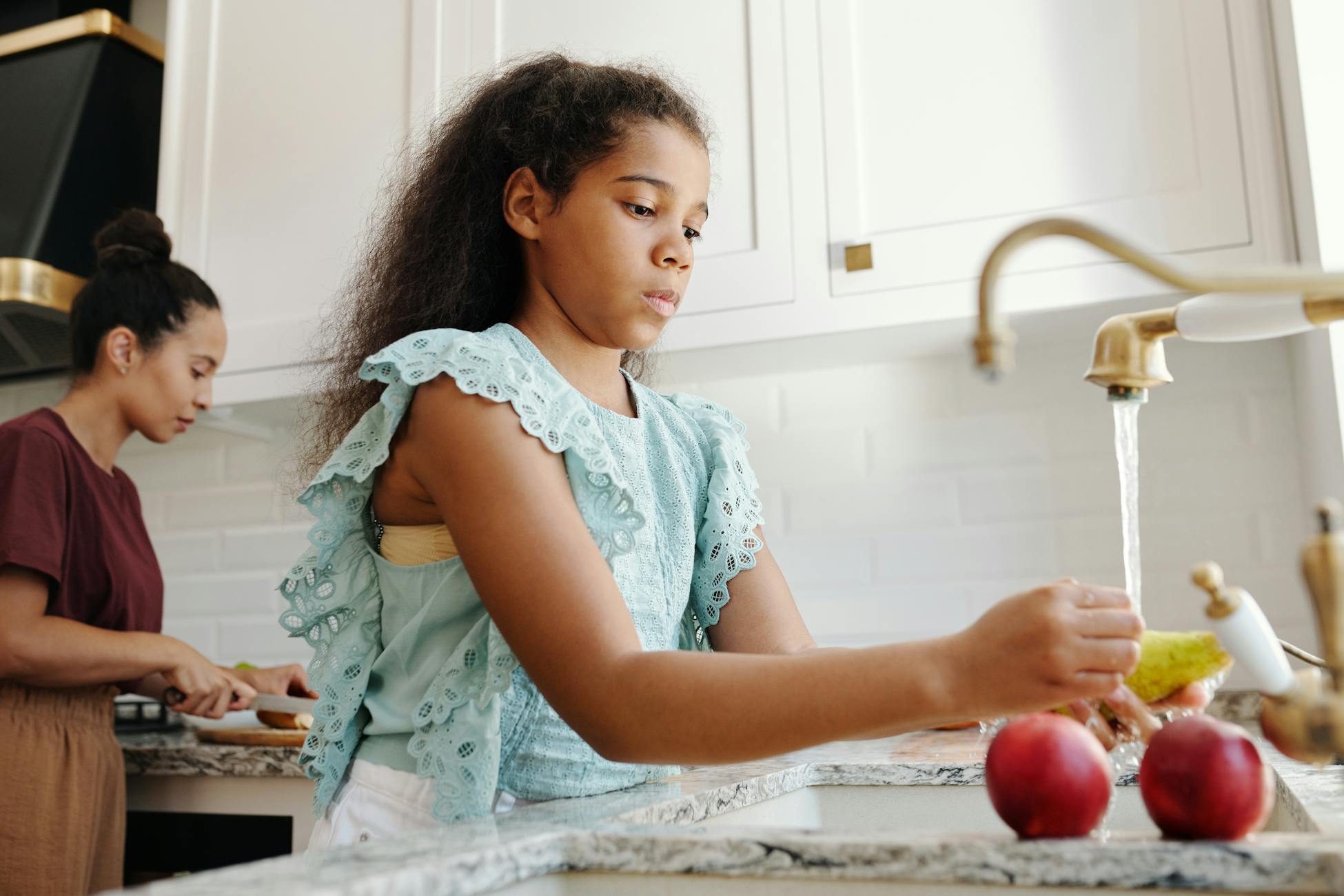 African American girl washing fruits at kitchen sink beside adult in elegant kitchen.