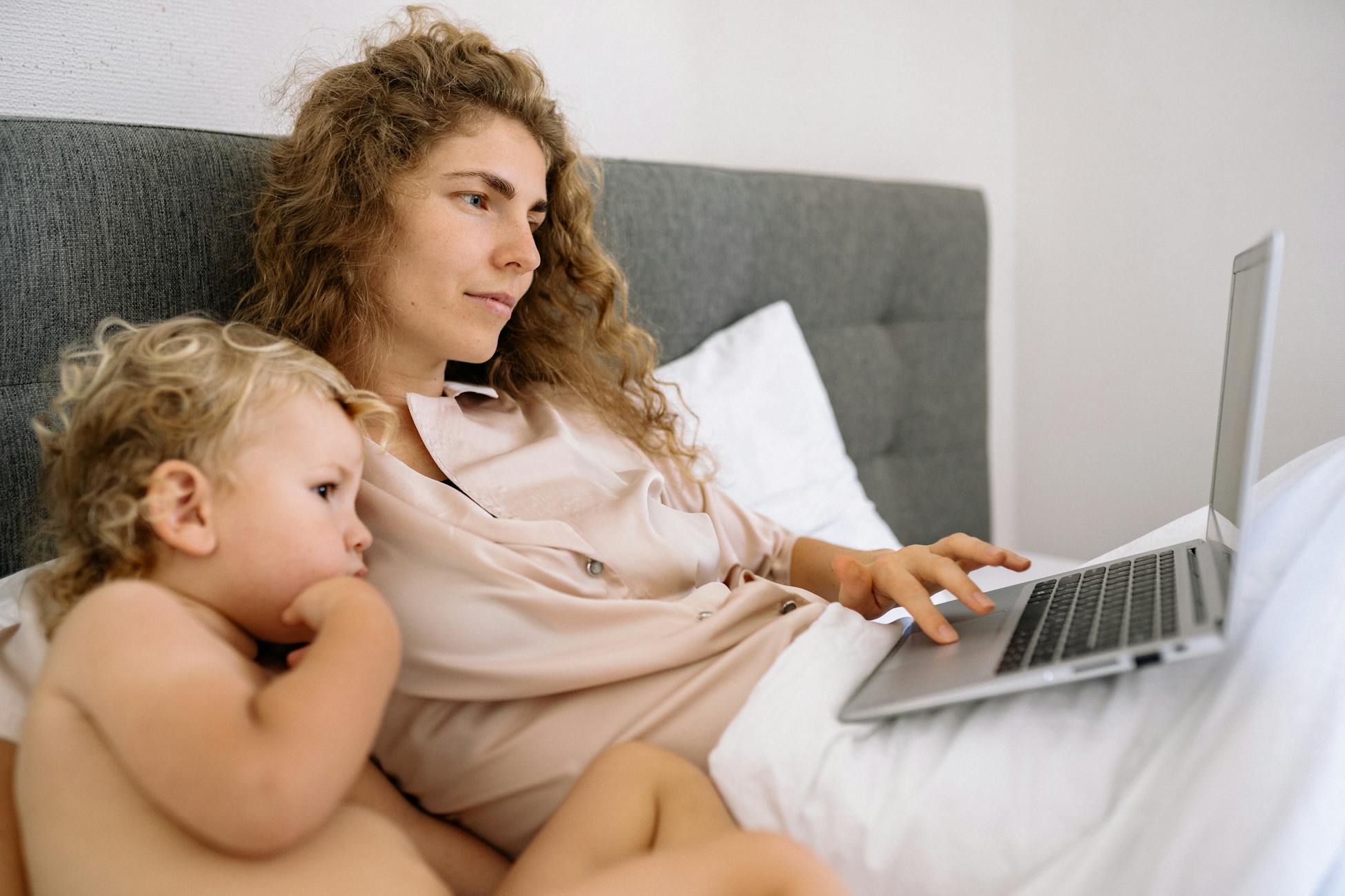 A mother and child bonding while watching content on a laptop in a cozy bed setting.