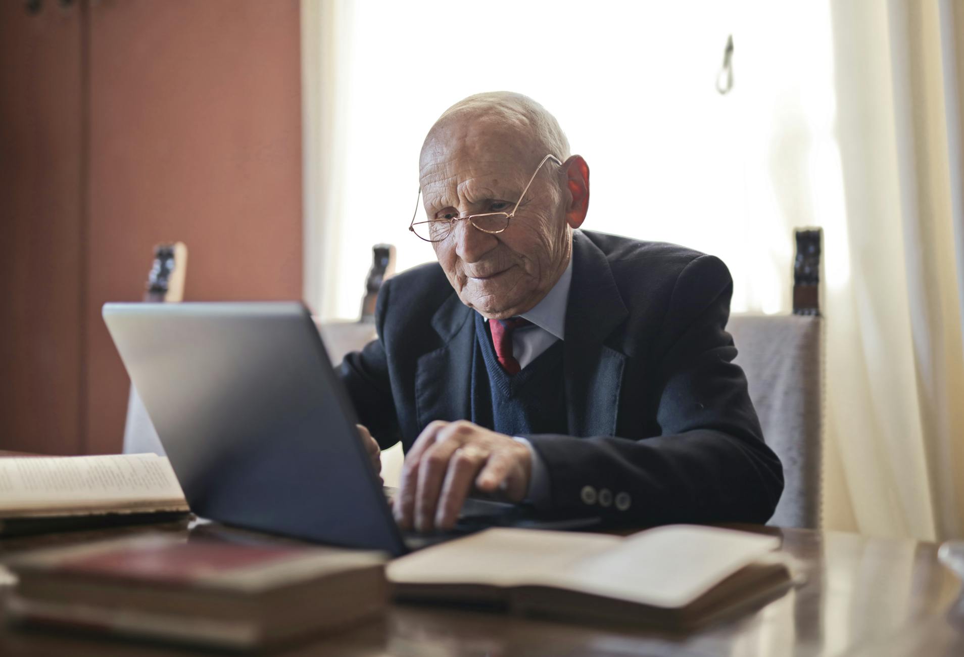 Focused elderly man in formal black suit and eyeglasses using laptop while sitting at wooden table with books in light room