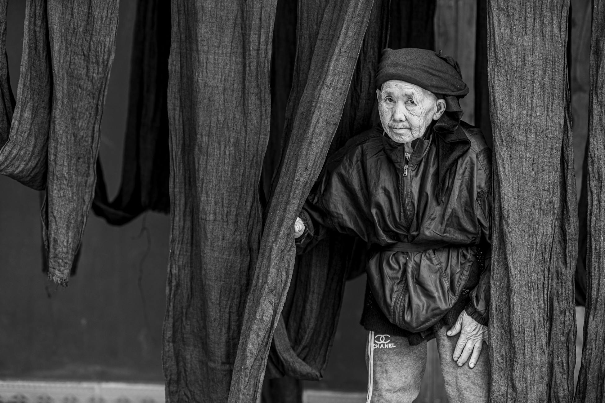 Monochrome portrait of elderly woman amidst draped fabric, deep in thought.