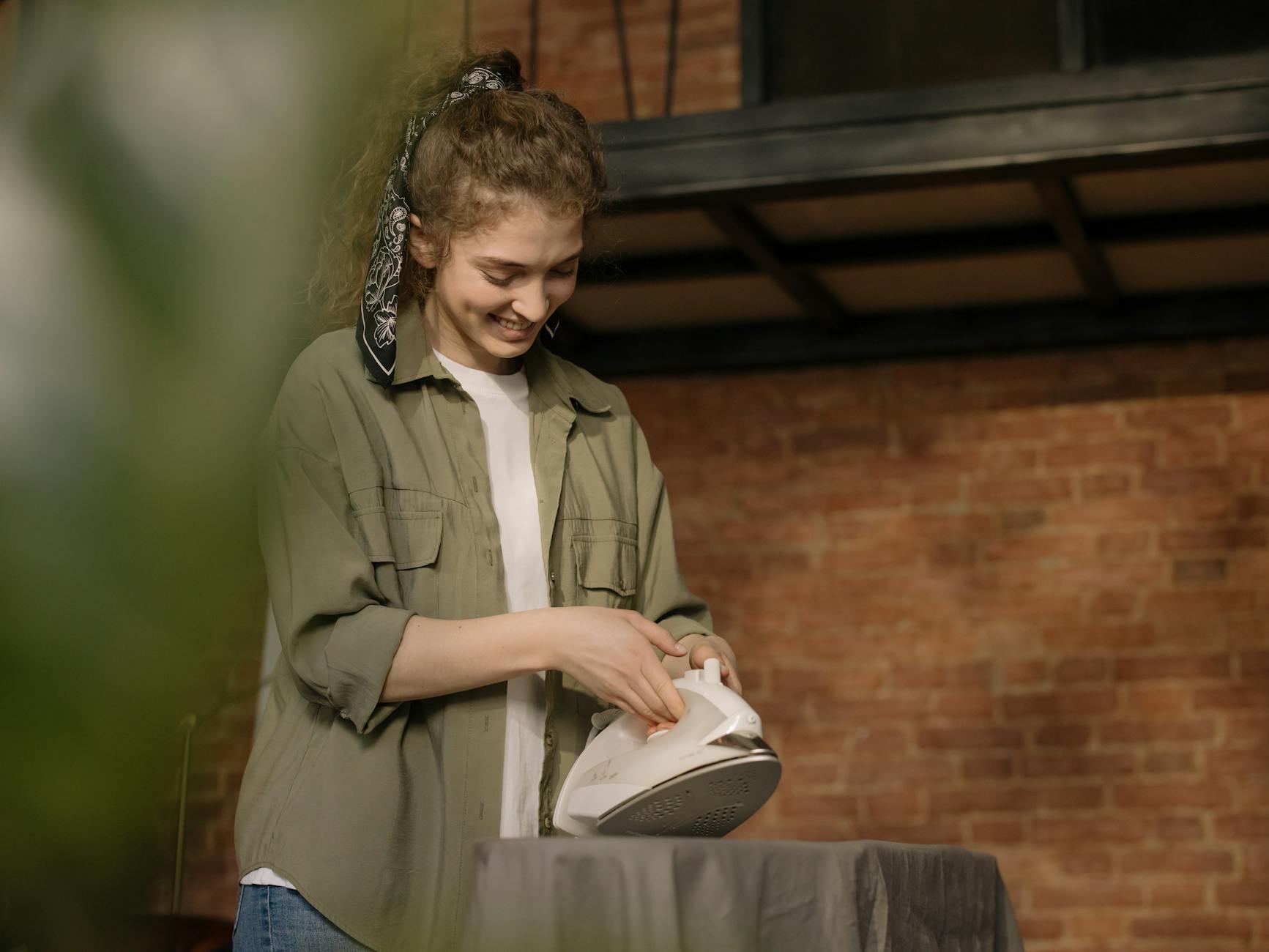 A young woman with curly hair ironing clothes in a cozy, modern home interior with a brick wall.