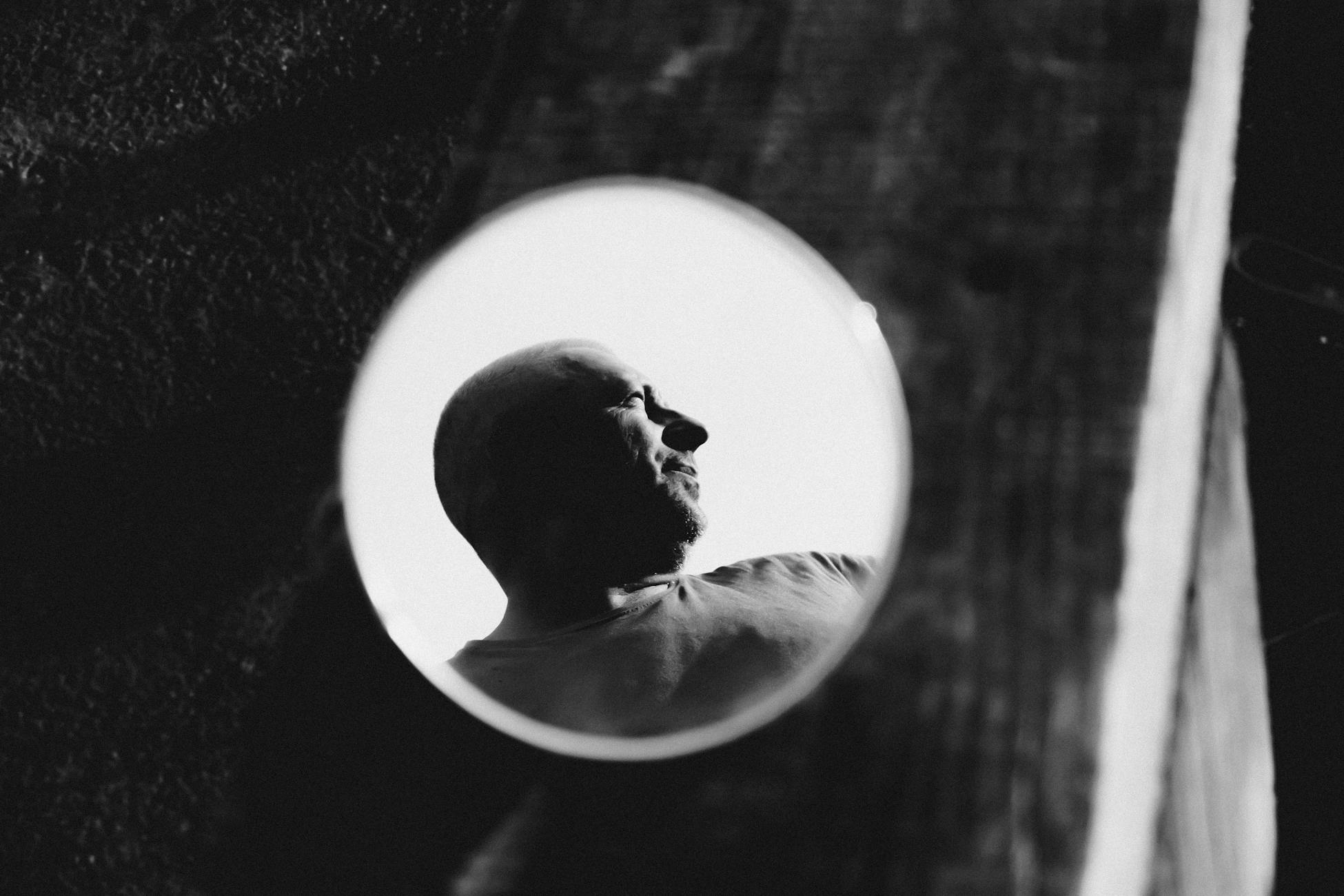 Black and white of hairless man reflecting in round mirror placed on dark background