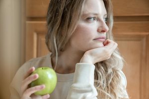 A young woman sitting indoors, pensively holding a green apple, gazing into the distance.