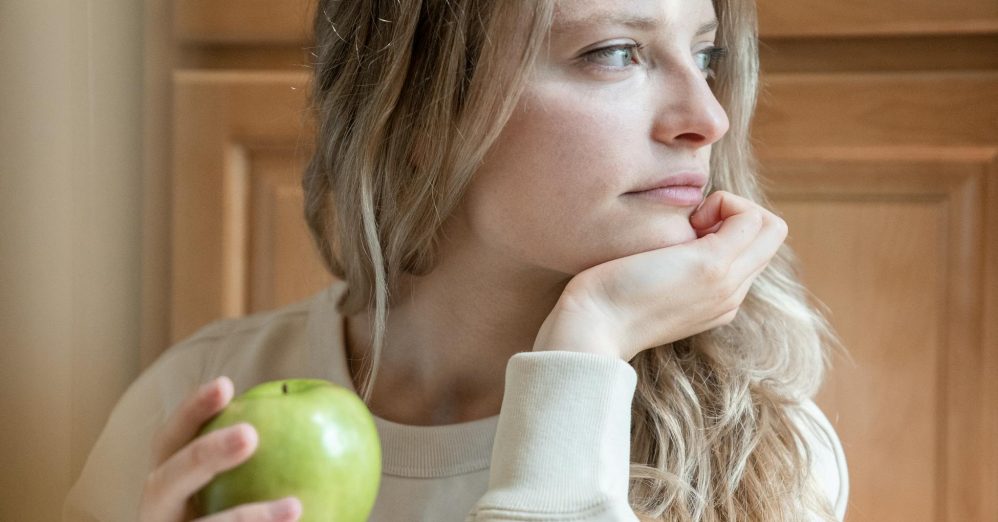 A young woman sitting indoors, pensively holding a green apple, gazing into the distance.
