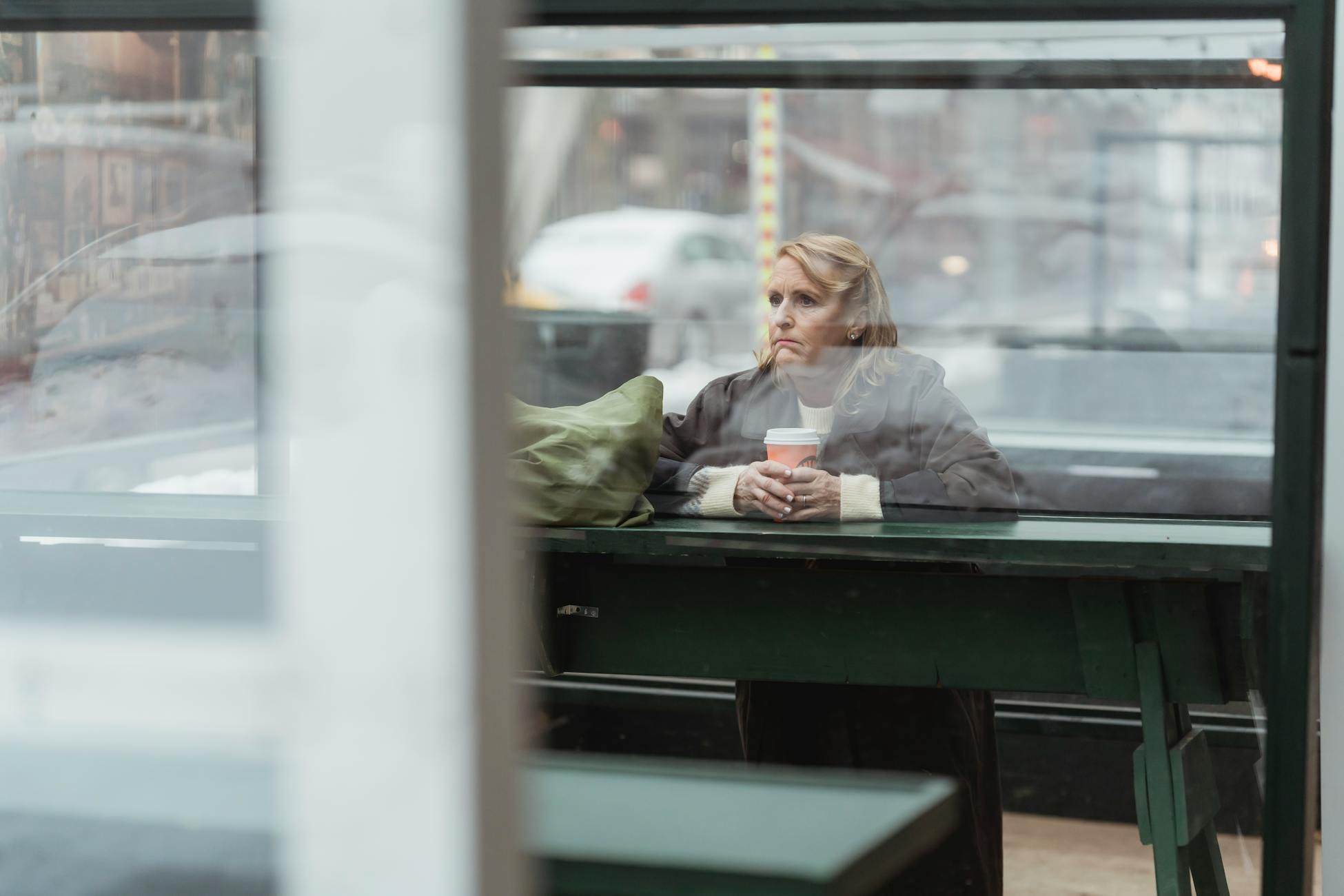 Pensive senior woman holding coffee in an outdoor café, reflecting through glass.