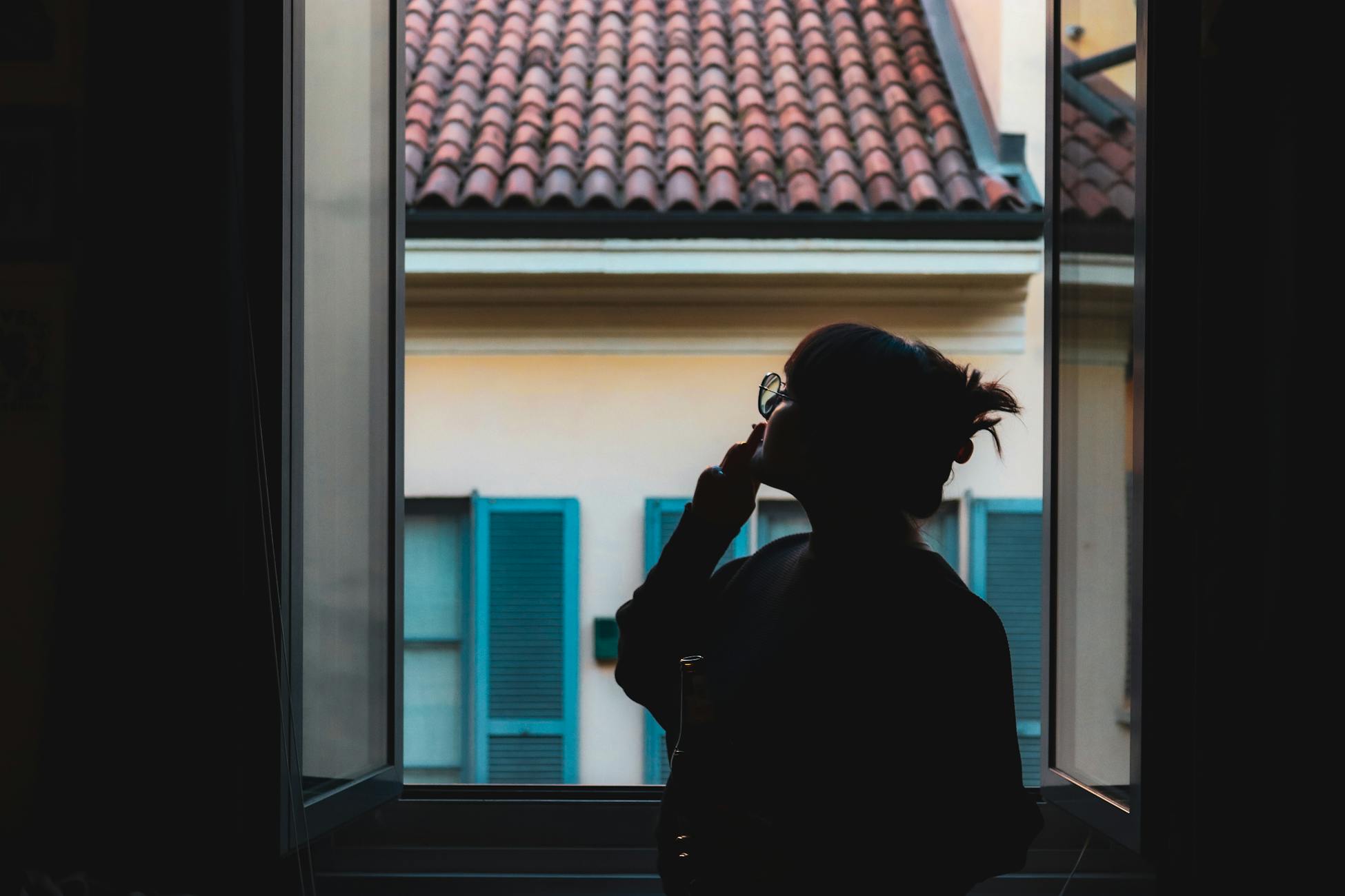 Silhouette of a woman in a serene moment by the window, viewing the tiled roof outside.