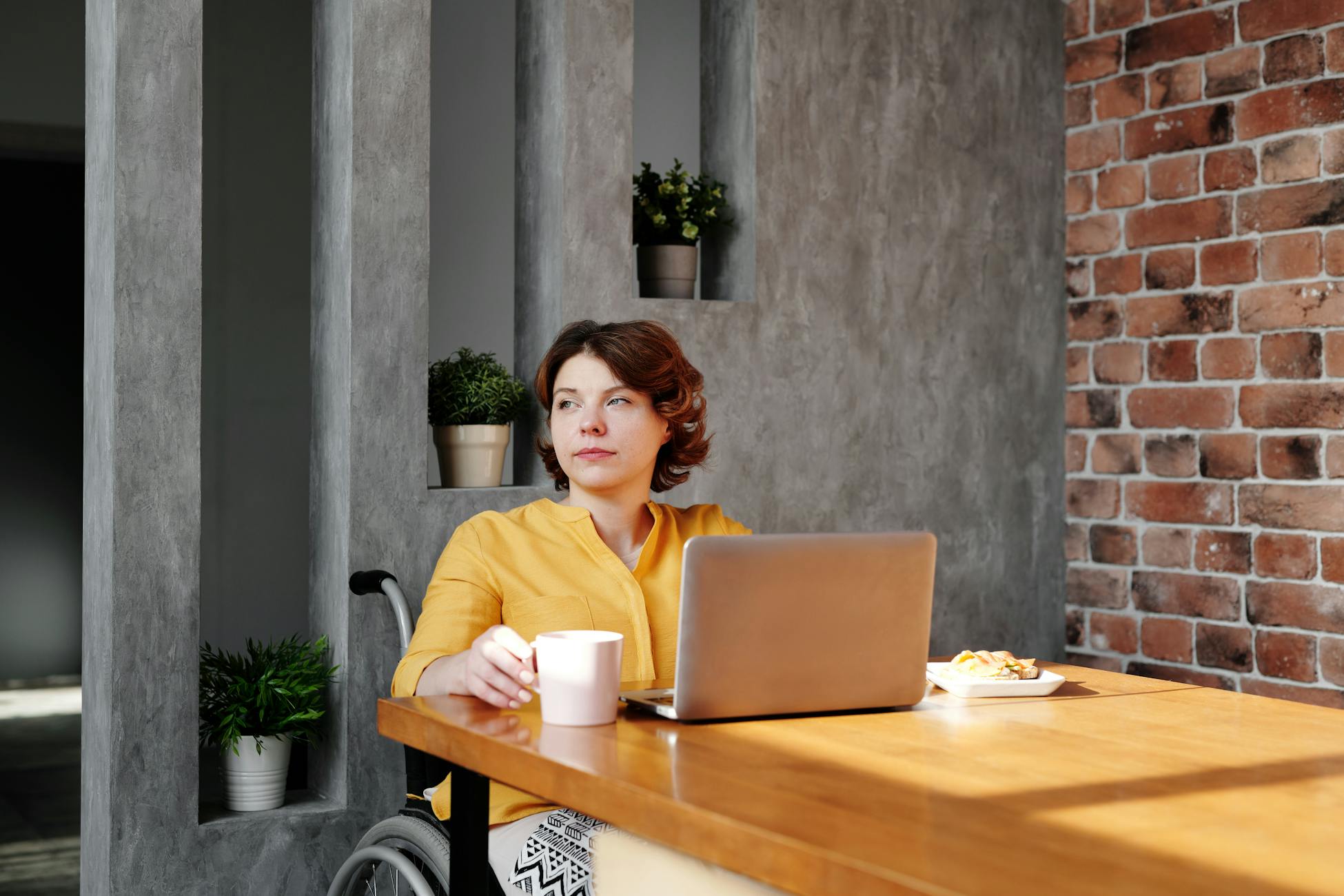A woman in a wheelchair working on a laptop at home, drinking coffee.