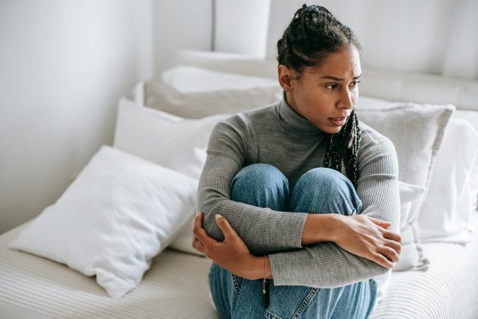 A woman sits thoughtfully on a bed, wrapped in her own arms, displaying signs of deep contemplation.
