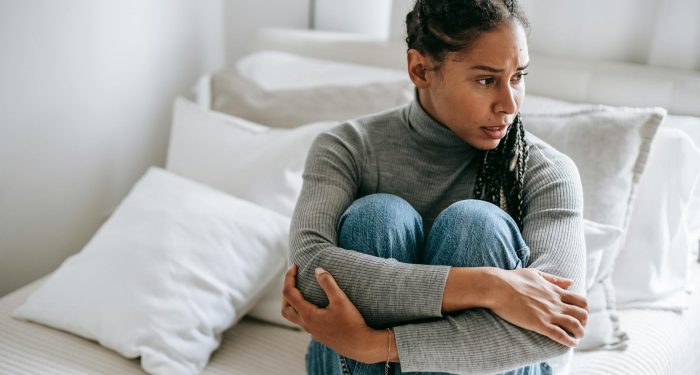 A woman sits thoughtfully on a bed, wrapped in her own arms, displaying signs of deep contemplation.