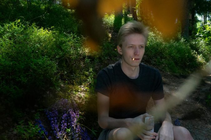 Pensive male in casual clothes smoking cigarette while sitting in forest in sunny summer day