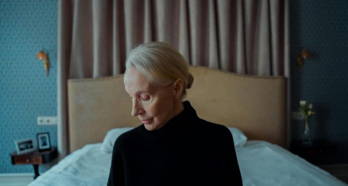 A senior woman reflects alone in a softly lit bedroom, capturing solitude.