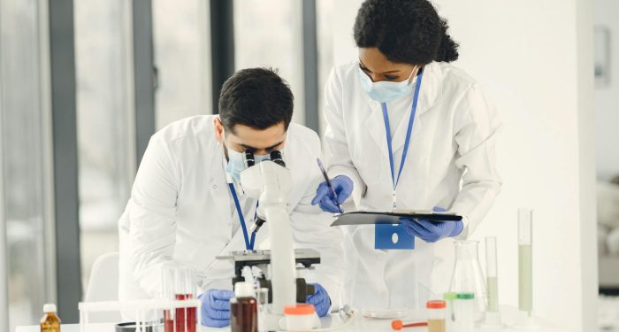 Researchers in lab coats and masks working with microscope and test tubes in a laboratory setting.