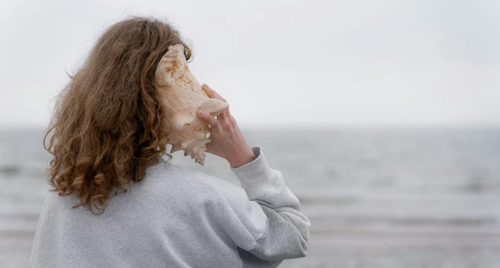 A young woman listens to a seashell by the ocean. Peaceful coastal scene.