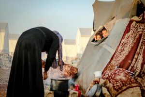 A mother cooks outside a tent while children peek out in a Syrian refugee camp.