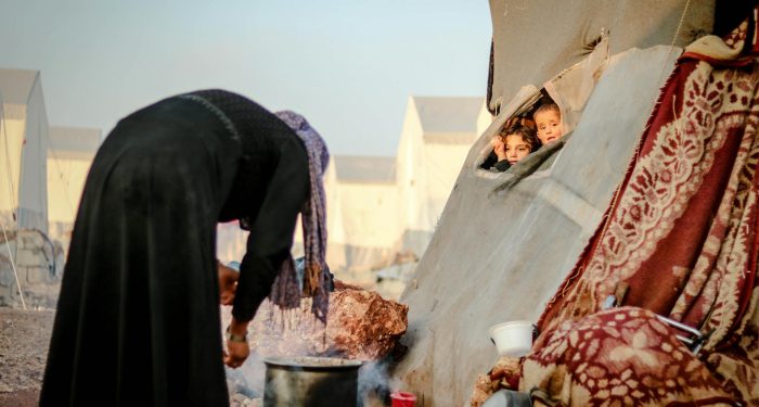 A mother cooks outside a tent while children peek out in a Syrian refugee camp.