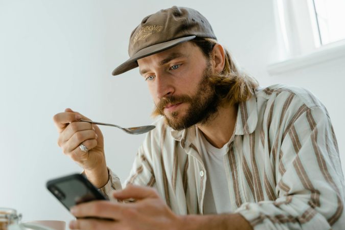 Bearded man wearing cap uses smartphone while eating at home.