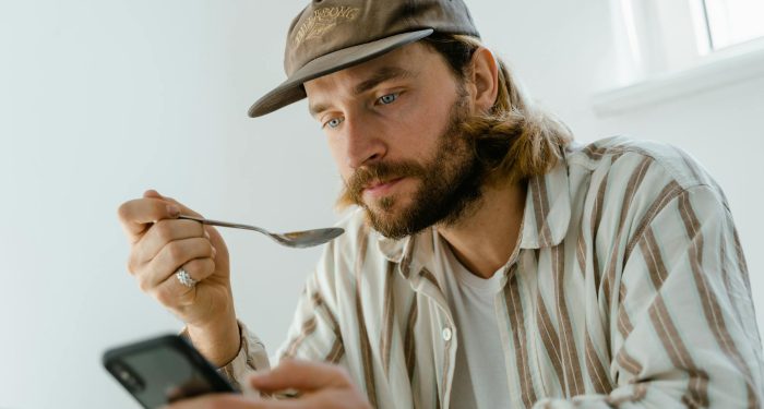 Bearded man wearing cap uses smartphone while eating at home.