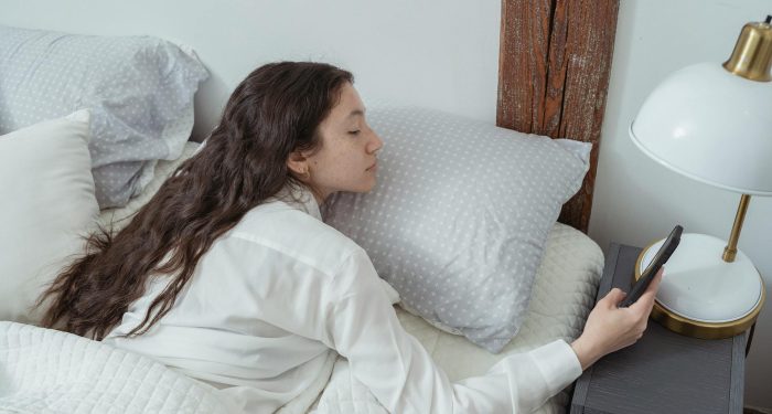 Side view of young brunette with long hair in white nightwear lying on belly in comfortable bed under blanket and using mobile phone in morning at home