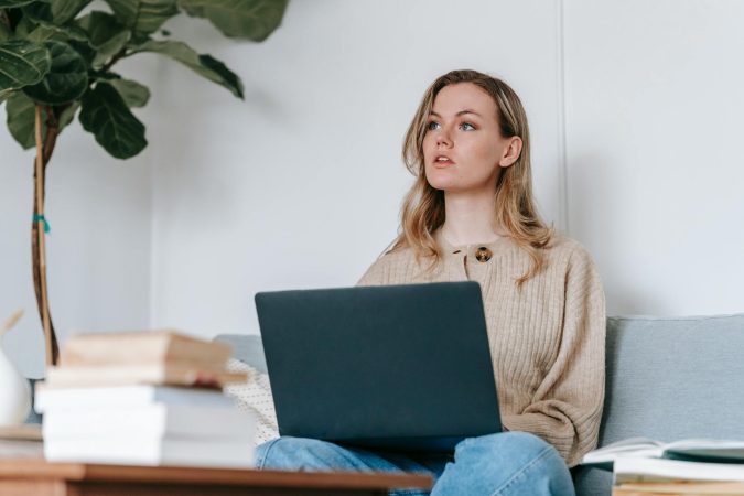 Young woman using a laptop while sitting on a couch, working remotely at home with books nearby.