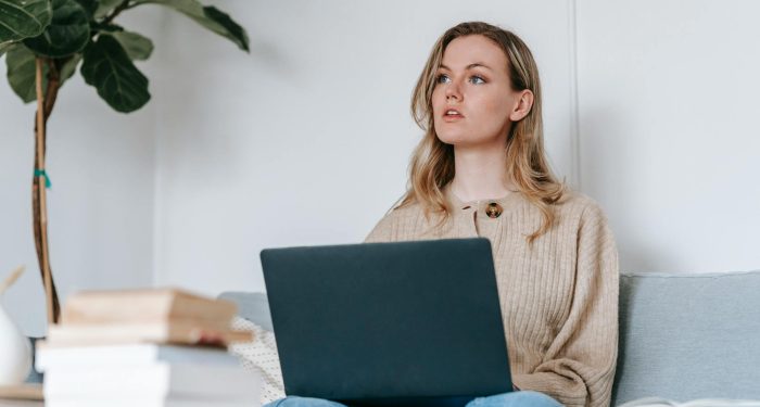 Young woman using a laptop while sitting on a couch, working remotely at home with books nearby.