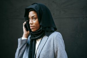 Serious young African American female in hijab and gray coat speaking on phone and looking away against black background