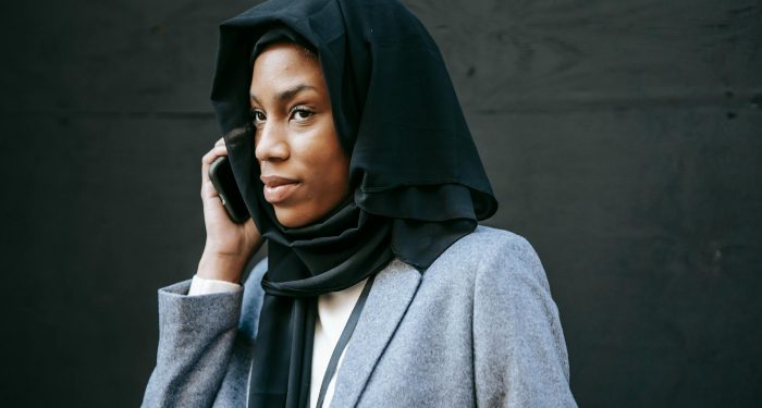 Serious young African American female in hijab and gray coat speaking on phone and looking away against black background