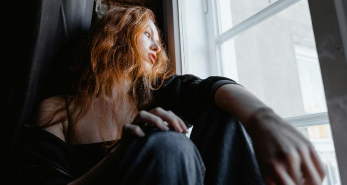 A woman with red hair sits by a window, deep in thought, conveying emotion and reflection.
