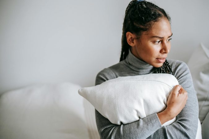 A woman in a grey sweater holds a pillow, looking contemplative in a serene indoor setting.