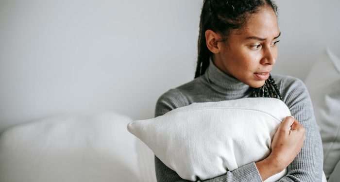 A woman in a grey sweater holds a pillow, looking contemplative in a serene indoor setting.