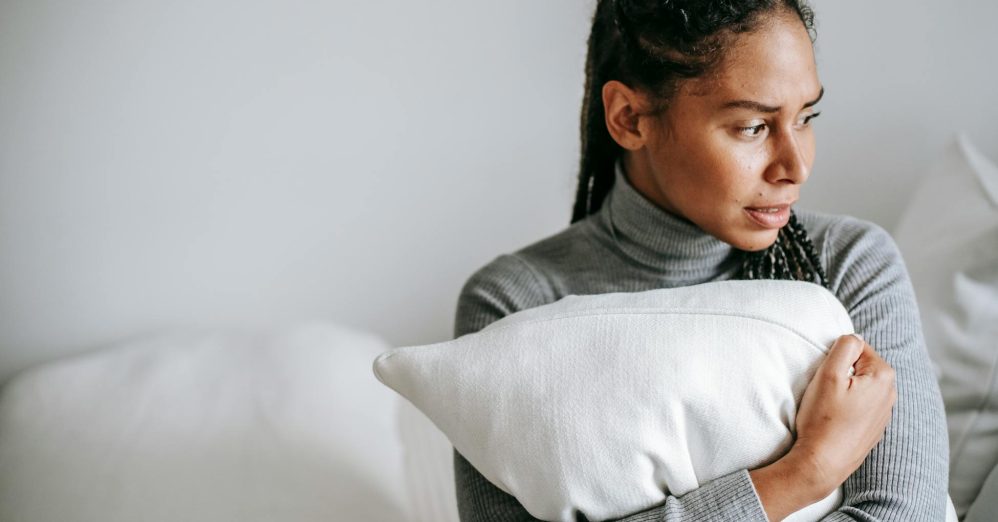 A woman in a grey sweater holds a pillow, looking contemplative in a serene indoor setting.