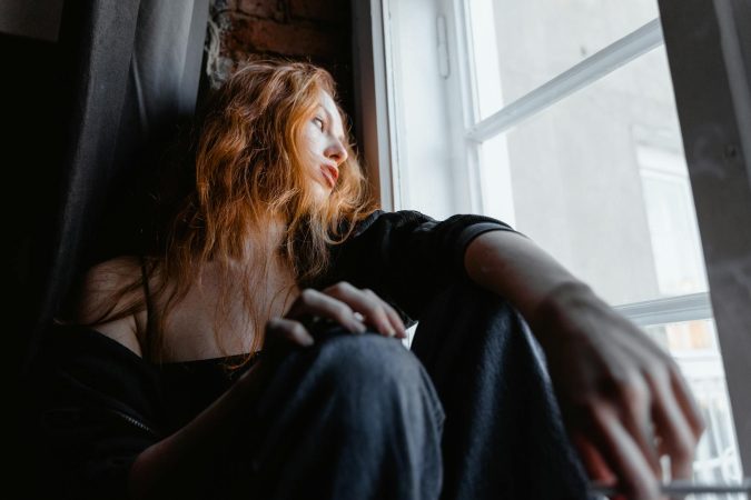 A woman with red hair sits by a window, deep in thought, conveying emotion and reflection.