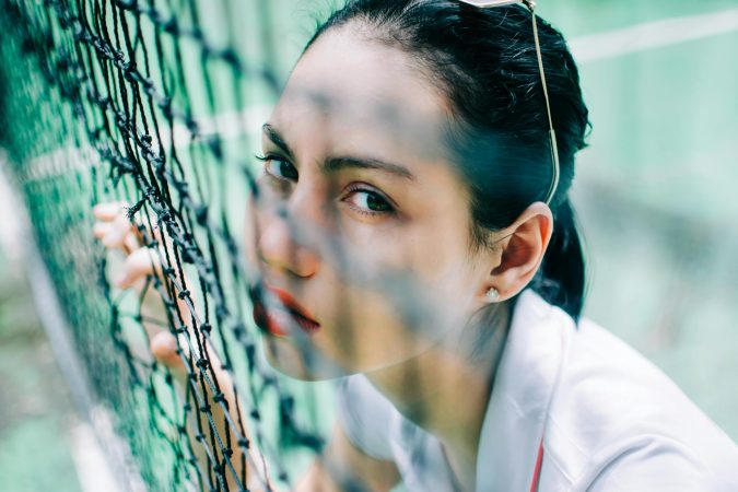 High angle of crop flirting female in t shirt looking at camera through net on tennis court