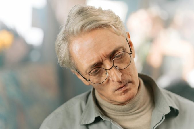Elderly man with glasses and gray hair appearing pensive and thoughtful indoors.