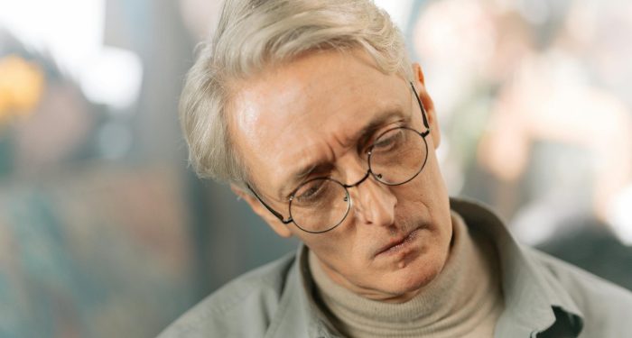Elderly man with glasses and gray hair appearing pensive and thoughtful indoors.