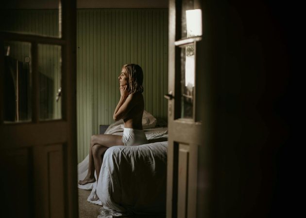 A woman sitting thoughtfully on a bed in a softly lit bedroom.