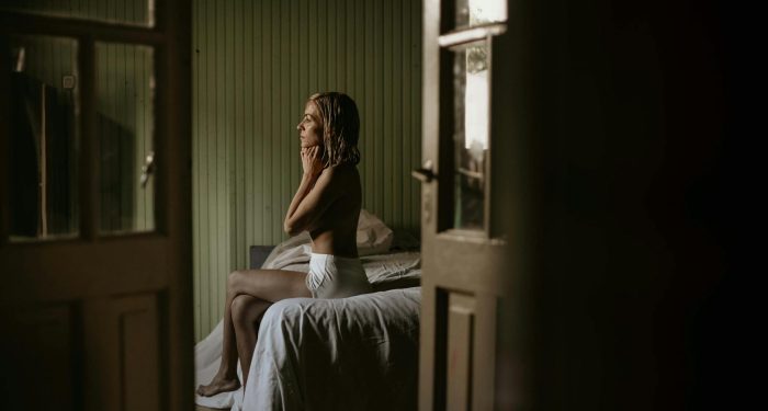 A woman sitting thoughtfully on a bed in a softly lit bedroom.