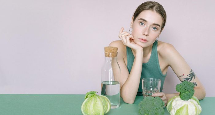 A young woman in a green top sitting with vegetables and a glass of water.