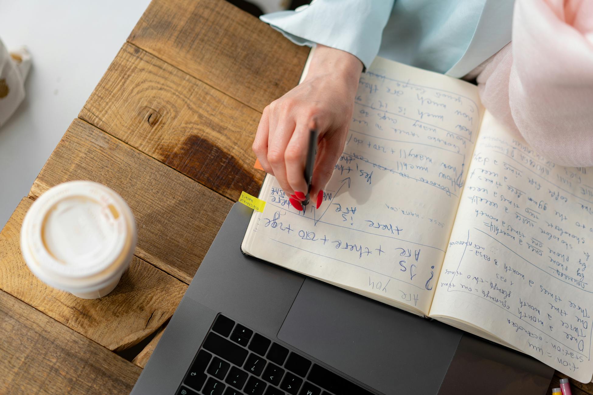 High-angle view of a woman taking notes in a planner with a coffee cup nearby on a wooden table.