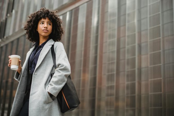Urban portrait of a confident woman in a gray coat holding coffee against a modern building.