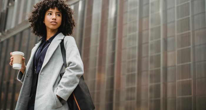 Urban portrait of a confident woman in a gray coat holding coffee against a modern building.