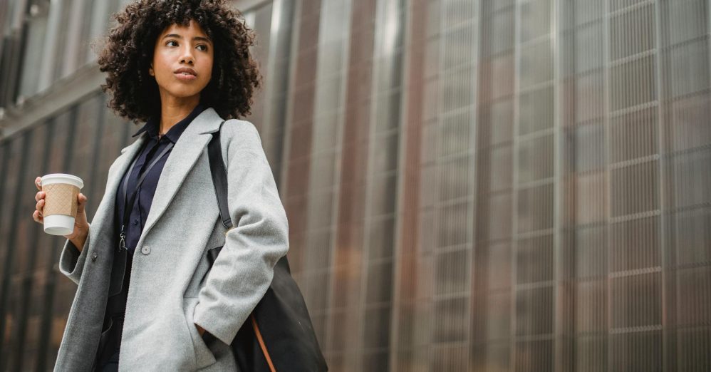 Urban portrait of a confident woman in a gray coat holding coffee against a modern building.