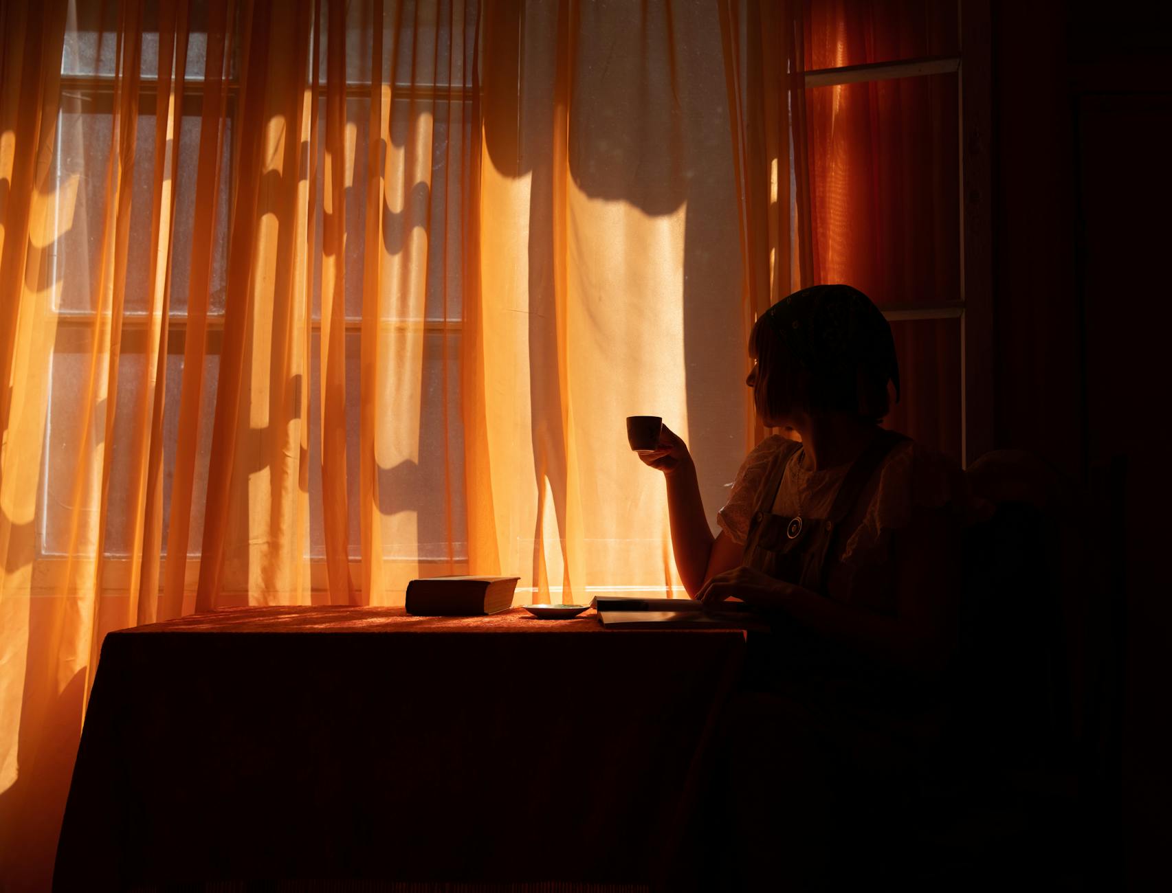 A woman enjoys a quiet moment with coffee, silhouetted by warm sunlight through curtains.