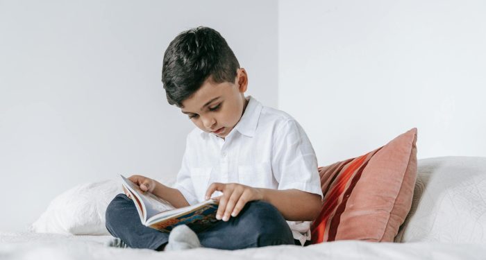 A young boy intently reading a book while sitting on a bed at home.