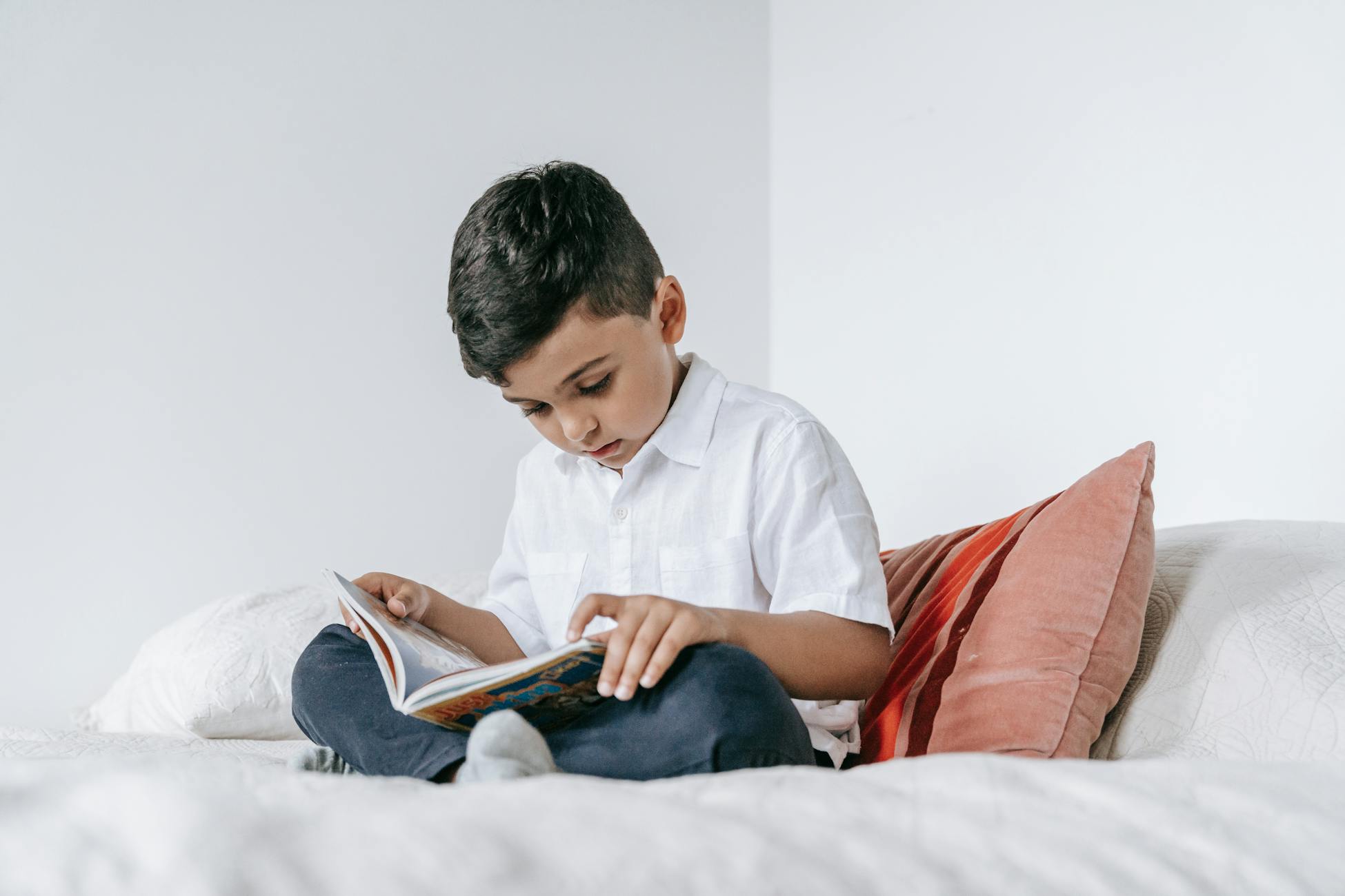 A young boy intently reading a book while sitting on a bed at home.