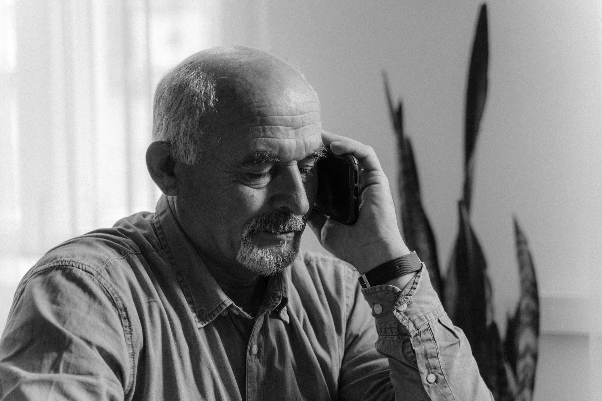 Elderly man engaged in a phone conversation indoors, captured in a grayscale portrait.
