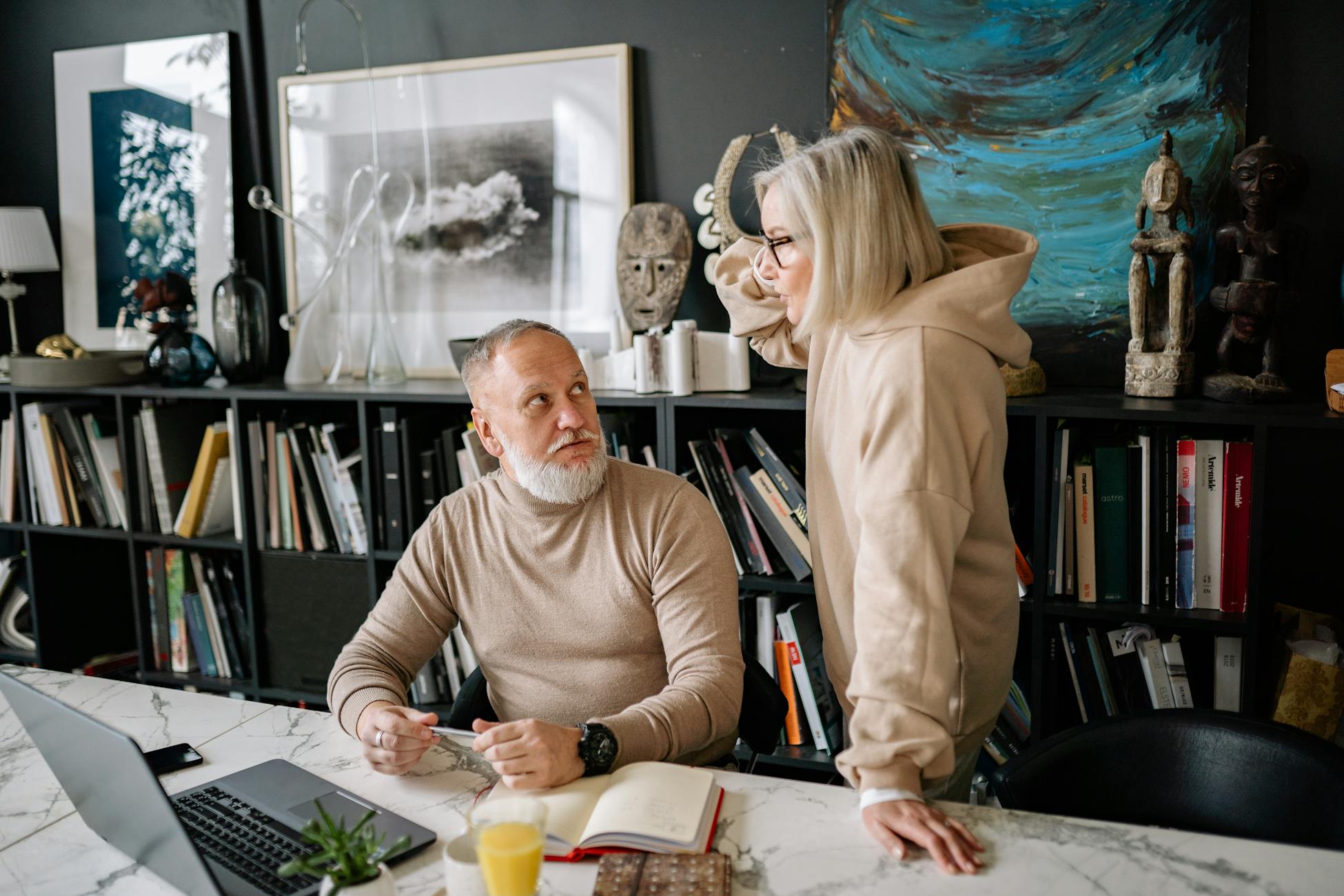 Elderly couple discussing something at a marble table with books and laptop, surrounded by art and sculptures.