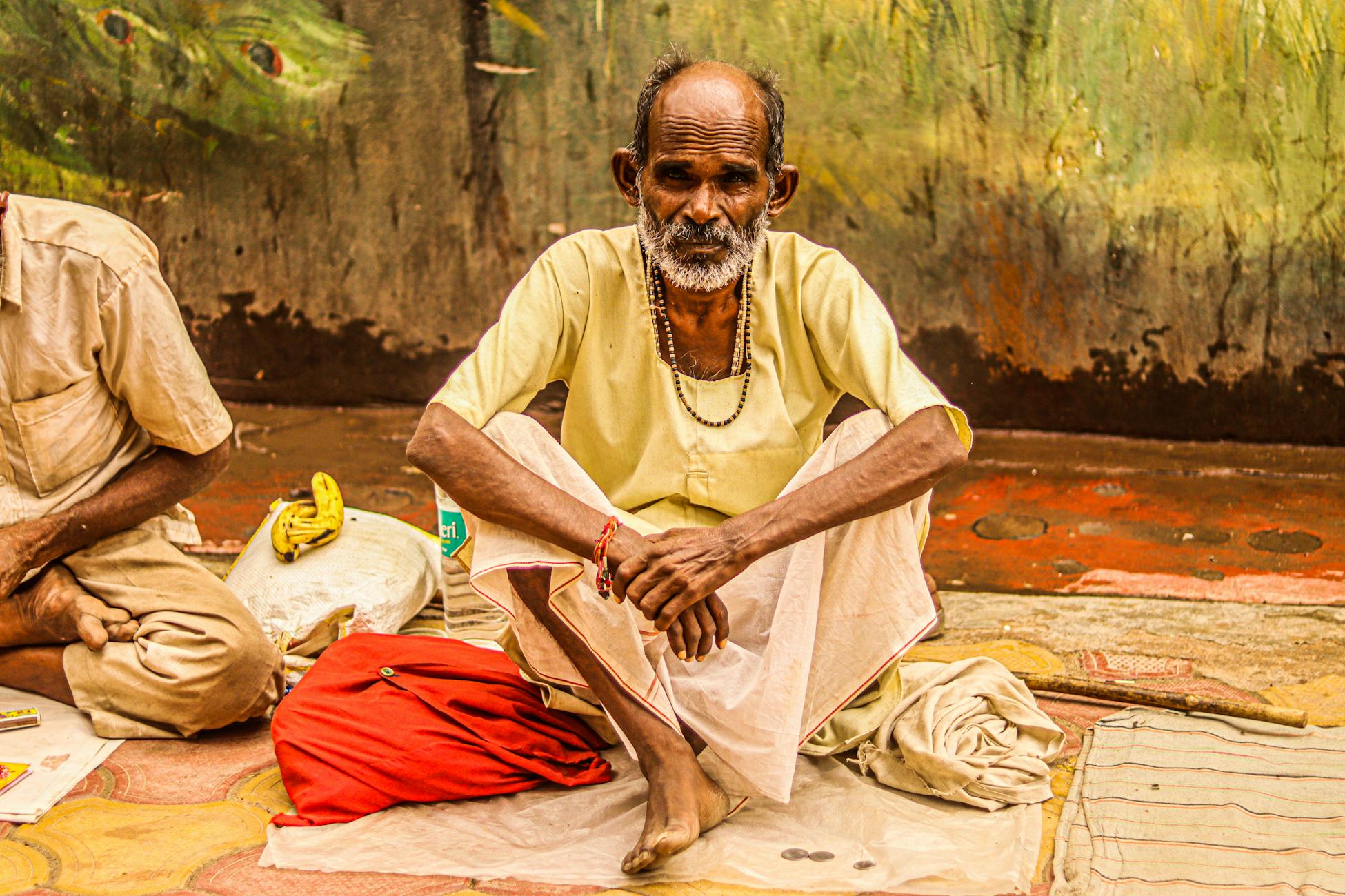 Elderly man sitting cross-legged, wearing a yellow shirt, outdoors.