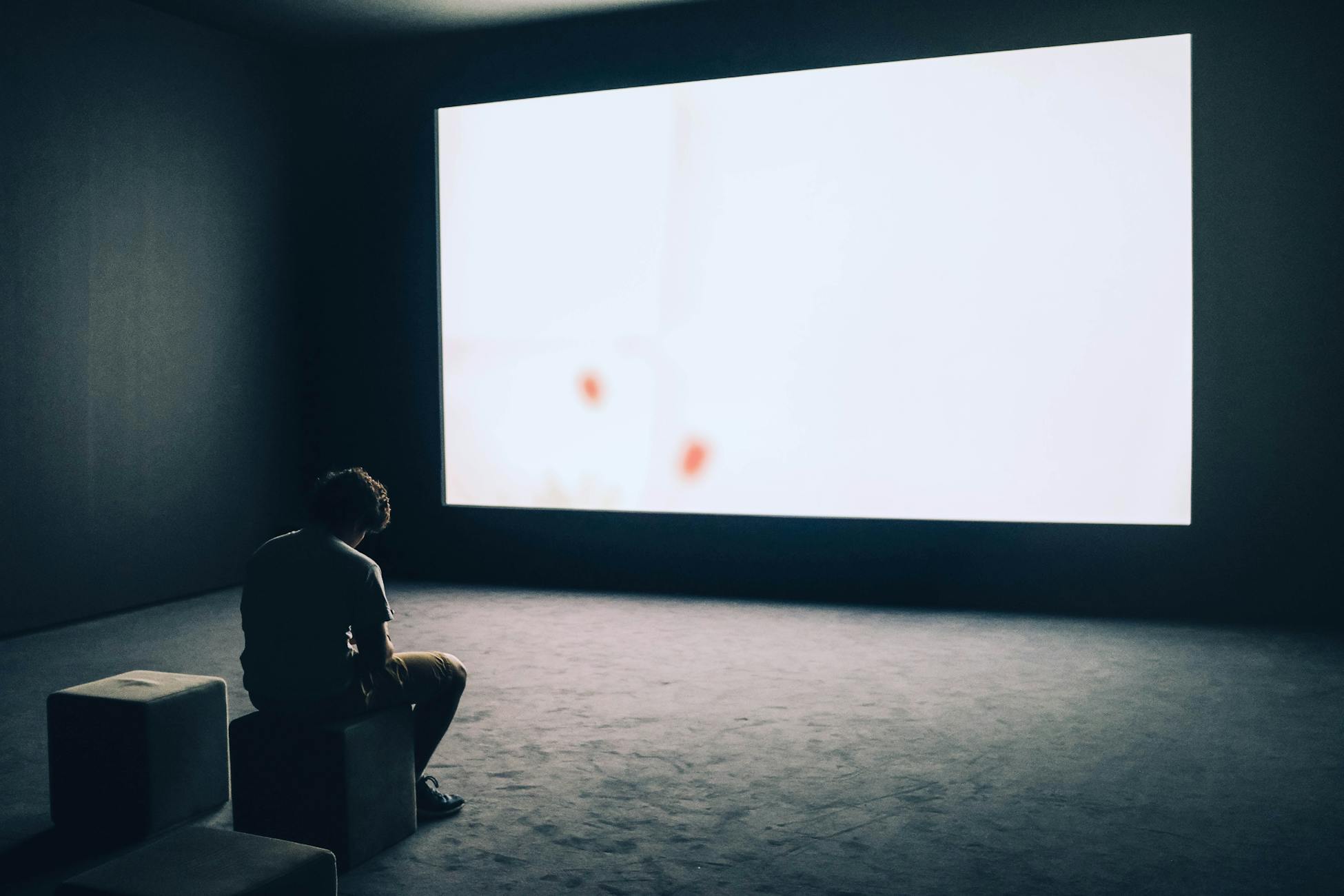 A man sits alone in a dark room with a large white screen, depicting solitude and contemplation.