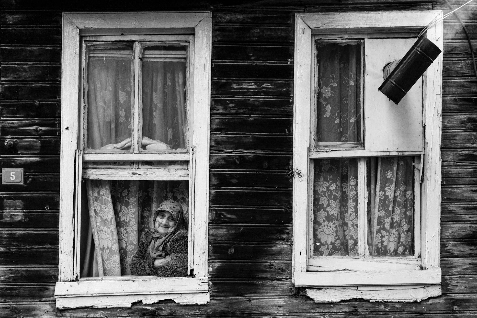 Black and white photo of an elderly woman looking out a vintage wooden window.